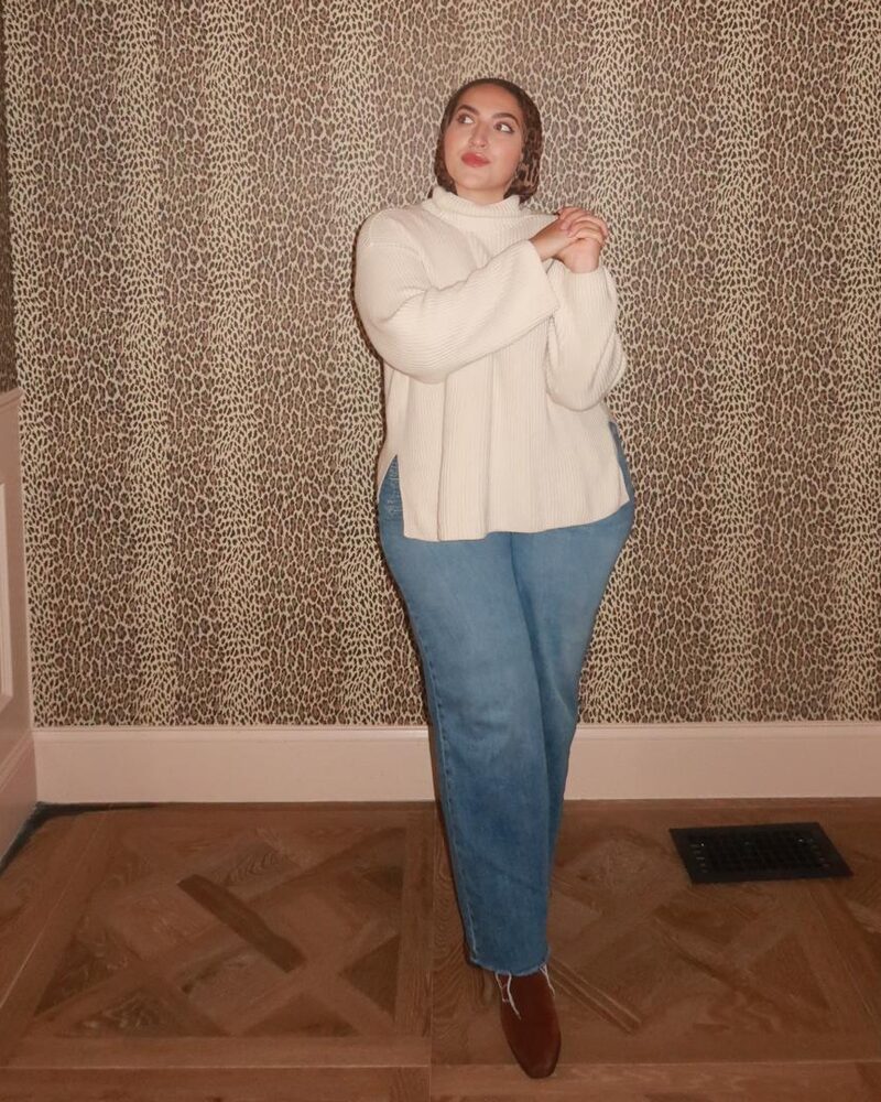 Naeema in a cream turtleneck sweater and jeans, hands clasped and looking up, against leopard-print wallpaper