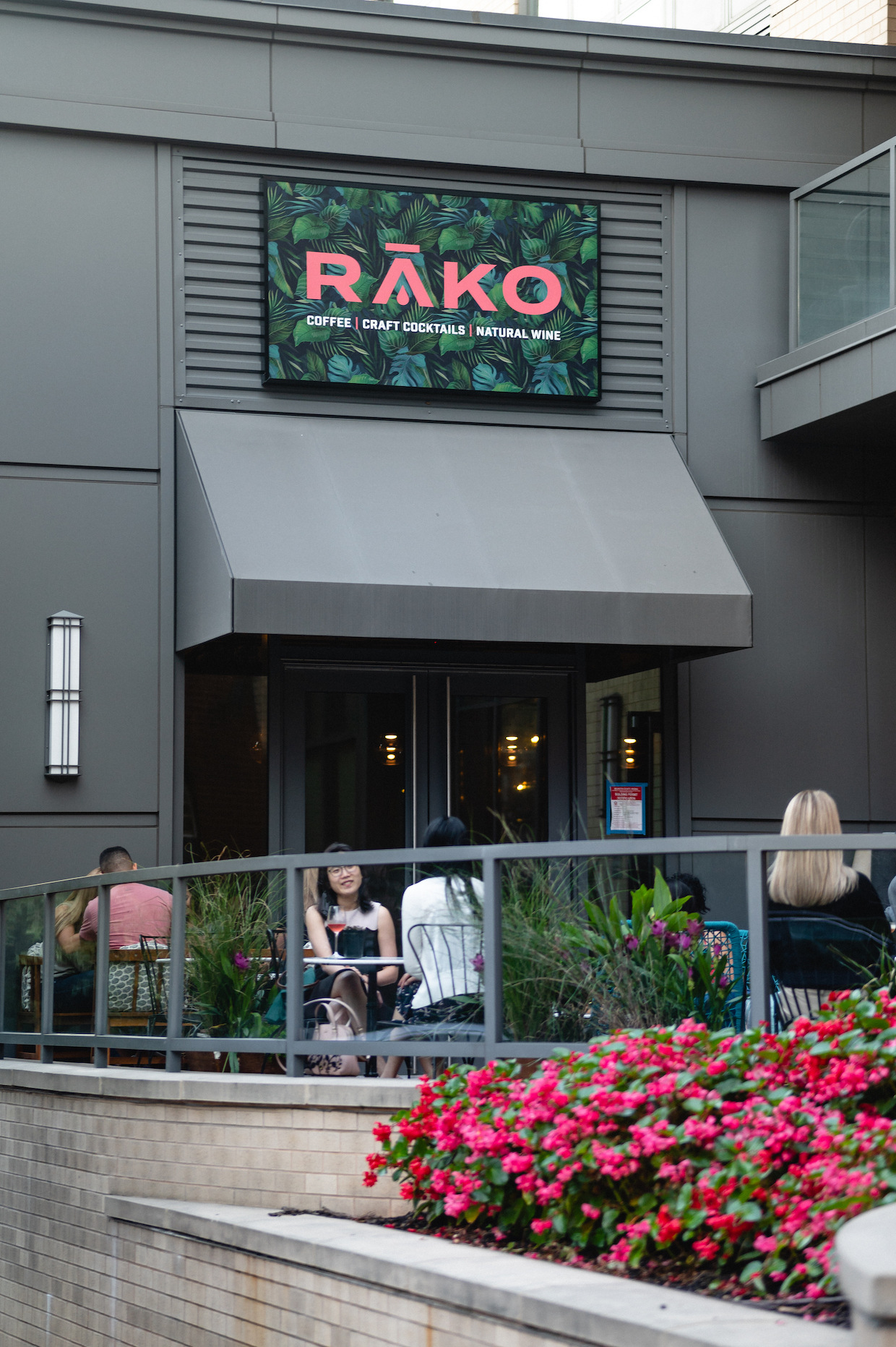 RĀKO's storefront with its pink logo on a tropical leaf sign, guests on the patio and pink flowers in front
